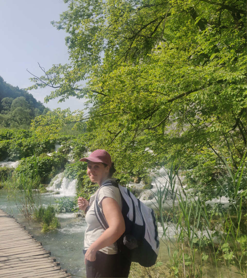 therapist jessica hughes hiking on a boardwalk next to a waterfall and greenery wearing a maroon hat and a backpack